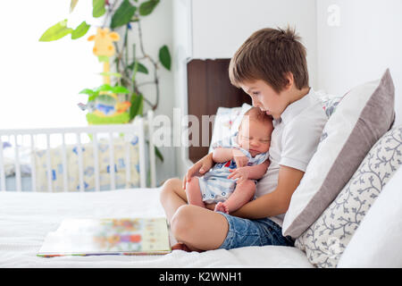 Sweet preschool boy, reading a book to his newborn brother, sitting on the bed in bedroom, childhood concept Stock Photo