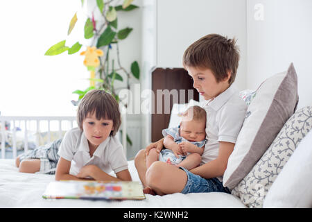 Sweet preschool boy, reading a book to his newborn brother, sitting on the bed in bedroom, childhood concept Stock Photo