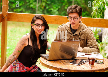 Handsome young man working in computer while her girlfriend is looking at camera, working on porch of wooden house, romantic moment, summer relaxing together concept, in a backyard background Stock Photo
