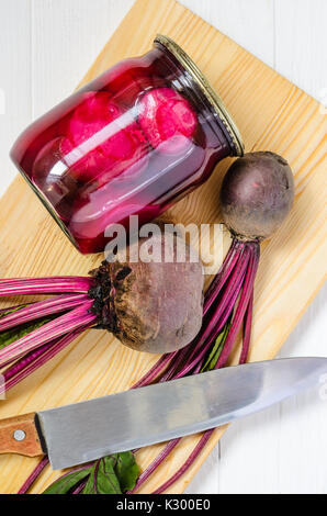 Glass jar with canned beets home made and fresh organic beets with haulm with cutting board and big knife on a white wooden background top view. Beets Stock Photo