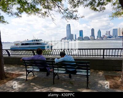 Two men sitting on a bench in New York's Battery Park watching boats pass by on the Hudson River with Jersey City, NJ in the background. New York, USA. Stock Photo