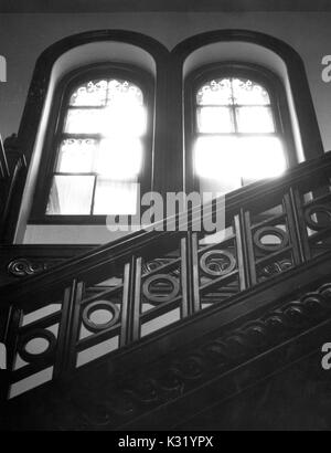 Grayscale photograph of wooden bannisters and two large windows with sun pouring in, from the interior of Clifton Mansion, built at the turn of the 18th century as a two story Federal farmhouse but later expanded into a Victorian era Italian villa, owned by philanthropist Johns Hopkins and later purchased by the City of Baltimore, Baltimore, Maryland, February, 1963. Stock Photo