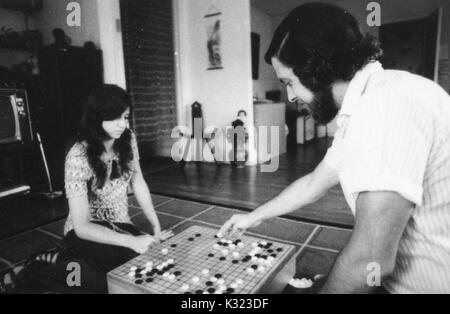 A male and female student sit on opposite sides of a grid board engaged in a game of Go, an old Asian strategy game, with white and black stones arranged on the grid, 1970. Stock Photo
