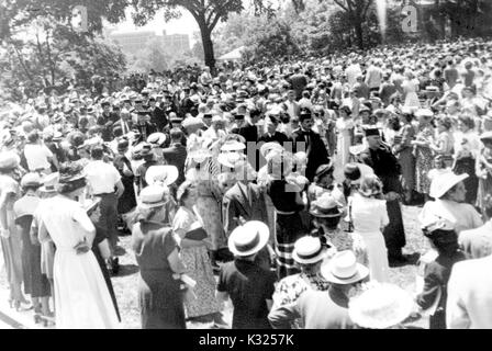 On Commencement Day, a large crowd of family, friends, and guests stand outdoors in clusters watching an aisle of graduates and faculty members proceed together wearing caps, gowns, and robes, Johns Hopkins University, Baltimore, Maryland, June, 1949. Stock Photo