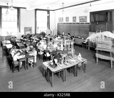 At an elementary demonstration school of Johns Hopkins University, students sit quietly at desks reading from workbooks while their teacher leads a reading group in front of the classroom beside the chalkboard, with a table of artifacts and objects on display off to the side of the room, Baltimore, Maryland, June, 1955. Stock Photo