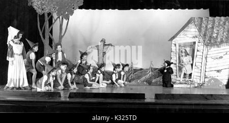 At an elementary demonstration school of Johns Hopkins University, boys and girls put on a performance of The Wizard of Oz on stage in an auditorium, several squatting on the stage floor wearing pointed witch hats, with a scarecrow, two witches, and the protagonist Dorothy, surrounded by a stage set consisting of a small house and trees, Baltimore, Maryland, June, 1947. Stock Photo