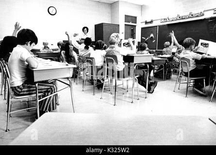At an elementary demonstration school for Johns Hopkins University, young boys and girls sit in desks in a classroom, while the female teacher stands at the side of the room, and several student raise their hands to answer a question, Baltimore, Maryland, June, 1975. Stock Photo