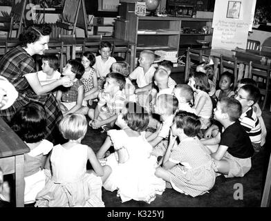 At an elementary demonstration school for Johns Hopkins University, students in Ms Catherine Brunner's second grade class sit around her in a semi-circle on the classroom floor, listening intently while she speaks to the students, smiling with her hands together, Baltimore, Maryland, June, 1955. Stock Photo