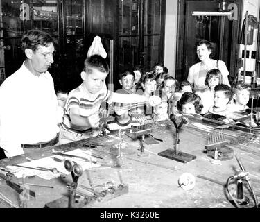 During a summer field trip to a glass blowing studio, young students at a Johns Hopkins University elementary demonstration school huddle around a table with their teacher, observing with fascination while a male classmate works a piece of glass tubing under the supervision of a glassblower beside him, July, 1962. Stock Photo