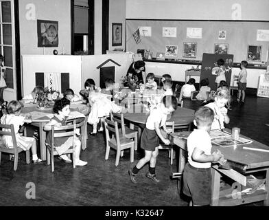 At an elementary demonstration school of Johns Hopkins University, students in Ms Berenice M Cronin's kindergarten class keep themselves busy while drawing, painting, telling stories, and sawing, as Ms. Cronin helps a student at a group table in the front of the classroom by the blackboard, Baltimore, Maryland, June, 1955. Stock Photo