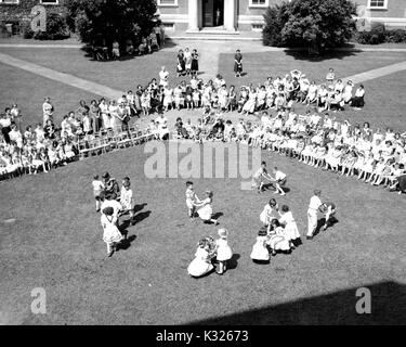 At the end of the school year for a demonstration school at Johns Hopkins University, young boys and girls put on a show in the grass on a sunny day, happily skipping and dancing in front of an audience made up of classmates, teachers, and parents sitting and standing outside of an ivy-covered campus building on an open quadrangle, Baltimore, Maryland, July, 1950. Stock Photo