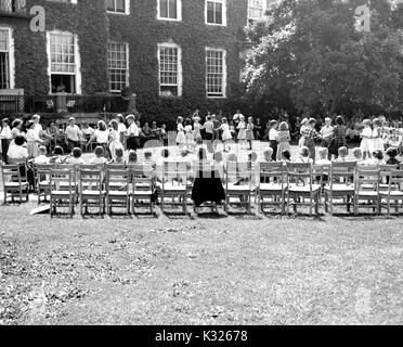 At the end of the school year for a demonstration school at Johns Hopkins University, young boys and girls put on a show in the grass on a sunny day, standing in circles holding hands in front of an audience made up of classmates and empty chairs lined up outside of an ivy-covered campus building, Baltimore, Maryland, July, 1950. Stock Photo