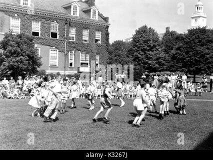 At the end of the school year for a demonstration school at Johns Hopkins University, young boys and girls put on a show in the grass on a sunny day, happily skipping in a circle in front of an audience made up of classmates, teachers, and parents sitting and standing outside of an ivy-covered campus building, Baltimore, Maryland, July, 1950. Stock Photo