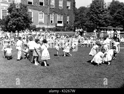 At the end of the school year for a demonstration school at Johns Hopkins University, young boys and girls put on a show in the grass on a sunny day, happily skipping and holding hands in front of an audience made up of classmates, teachers, and parents sitting and standing outside of an ivy-covered campus building, Baltimore, Maryland, June, 1950. Stock Photo