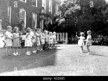 At the end of the school year for a demonstration school at Johns Hopkins University, young boys and girls participate in outdoor games on a sunny day, standing in a line while two girls hold batons and lead the group, in front of an audience made up of classmates, teachers, and parents sitting in the shade outside of an ivy-covered campus building, Baltimore, Maryland, July, 1950. Stock Photo