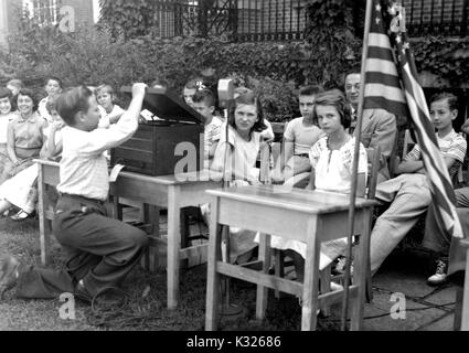 At the closing day activities for an elementary demonstration school at Johns Hopkins University, students sit at chairs in a row while one classmate prepares a music player and an American flag before for the singing of the national anthem, outside on the quadrangle in front of an ivy-covered campus building, Baltimore, Maryland, July, 1950. Stock Photo