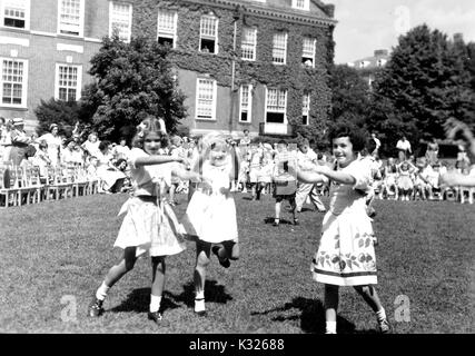At the end of the school year for a demonstration school at Johns Hopkins University, young boys and girls put on a show in the grass on a sunny day, happily skipping and waving their hands in front of an audience made up of classmates, teachers, and parents sitting and standing outside of an ivy-covered campus building, Baltimore, Maryland, July, 1950. Stock Photo