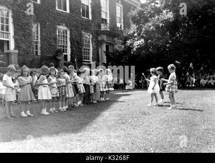 At the end of the school year for a demonstration school at Johns Hopkins University, young boys and girls participate in outdoor games on a sunny day, standing in a line while three girls hold batons and lead the group, in front of an audience made up of classmates, teachers, and parents sitting in the shade outside of an ivy-covered campus building, Baltimore, Maryland, July, 1950. Stock Photo