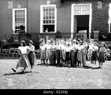 At the end of the school year for a demonstration school at Johns Hopkins University, boys and girls put on a show in the grass on a sunny day, standing in a line behind a microphone while two girls wearing skirts show off their dance moves, outside of an ivy-covered campus building, Baltimore, Maryland, July, 1950. Stock Photo