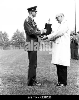 On the grassy Homewood Field at Johns Hopkins University, President Milton Stover Eisenhower wears a trench coat and glasses while presenting the Presidents Trophy to ROTC Cadet Alexander Davidoff, dressed in uniform, looking proud, with a line of other cadets to the side, Baltimore, Maryland, 1967. Stock Photo