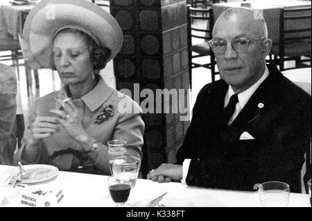During the Associazione Italo-Americana Luncheon at the School of Advanced International Studies (SAIS) in Washington, DC, President of Johns Hopkins University Milton Stover Eisenhower -- around 62 years of age -- sits at a table with his wife who smokes a cigarette, wearing a suit while his wife wears a blazer and hat, with wine glasses and water cups on the table, Washington, DC, 1965. Stock Photo