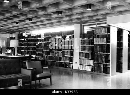 On an underground reading and study level inside the Milton S Eisenhower Library at Johns Hopkins University, stacks of books stand next to tables and lamps for studying, with one student hunched over a book in the background, in a quiet, dimly lit space, Baltimore, Maryland, 1965. Stock Photo