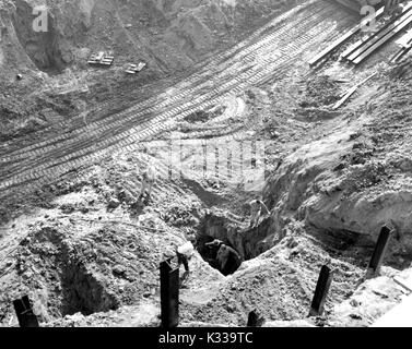 During the early stages of construction of the Milton S Eisenhower Library at Johns Hopkins University, workers wearing uniforms dig holes into the dirty to create a foundation for the building, with steel beams rising out of the ground, and the tracks of construction vehicles visible in the dirt, Baltimore, Maryland, 1962. Stock Photo