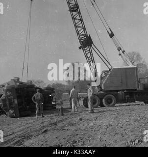 During the early stages of construction of the Milton S Eisenhower Library at Johns Hopkins University, an overturned drunk is lifted back to its upright position by a large crane, while three workers in boots and uniforms stand to oversee the process, above a rocky and muddy ground, Baltimore, Maryland, 1963. Stock Photo