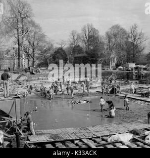 During the early stages of construction of the Milton S Eisenhower Library at Johns Hopkins University, cement is poured into the foundation of the building, with dozens of workers busy on site, and trees and campus buildings bordering the project ground, Baltimore, Maryland, 1962. Stock Photo