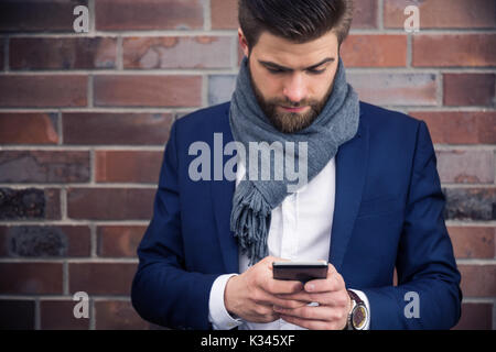A photo of young man in navy suit and shawl, standing in front of brick wall. He's using mobile phone. Stock Photo