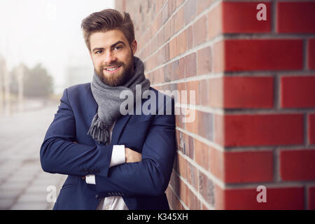 A photo of young man in navy suit and shawl, leaning against brick wall. He's smiling confidentnly. Stock Photo