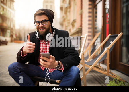 A photo of young man sitting on the deckchair with mobile phone in his hand. He's showing the thumb up. Stock Photo