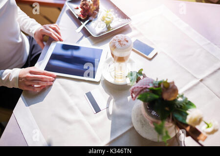 A photo of cafe table with glass of cappuccino, plate with cake, some decorations and electronic gadgets on it. Stock Photo