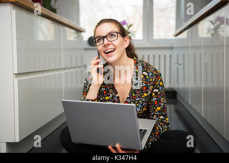 A photo of young woman sitting on the floor in the kitchen and talking on the mobile. She's wearing flowered shirt and holding laptop. Stock Photo