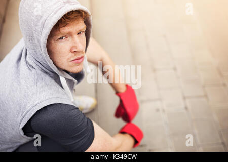 A photo of young man in red boxing gloves and a hood on his head. His sitting on the stairs. Stock Photo