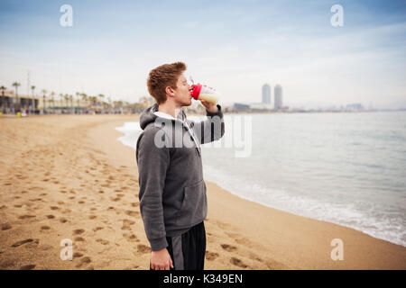 A photo of young sportsman standing on the beach and drinking electrolytes. Stock Photo