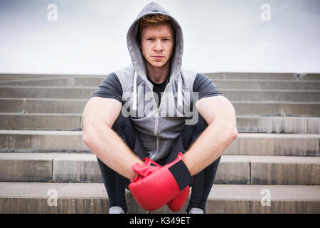 A photo of young boxer sitting on the city stairs. He's wearing a hood and red boxing gloves. Stock Photo