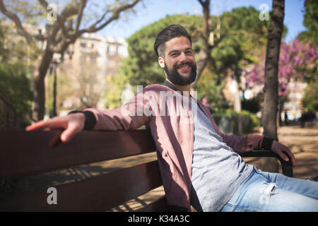 A photo of young man sitting on the park bench and smiling happily. He's enjoying sunny afternoon. Stock Photo