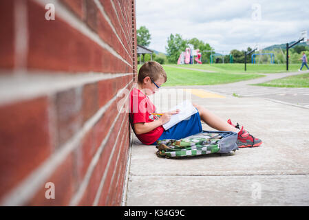 A boy studies a book while leaning against a brick wall of a school Stock Photo