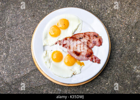 A traditional English breakfast of two eggs (with double yolks) and bacon on a white plate against a stone or granite background Stock Photo