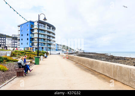 Man and woman seated on a bench facing the sea wall on the promenade at Westward Ho!, Devon, England, UK, holiday apartments in the background Stock Photo