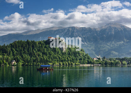 Traditional Pletna boat rowing tourists on Lake Bled with Bled castle on cliff and St Martin church Sol massive of Karawanks mountains Slovenia Stock Photo