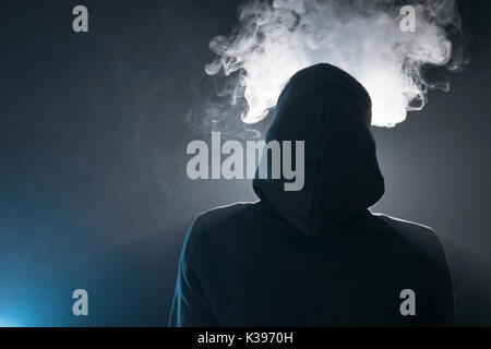 Isolated young man smoking an electronic cigarette on a dark background, holding a vaping device with lots of clouds. Stock Photo