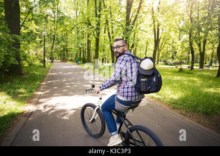 Rear portrait of young modern man riding bike at park with backpack Stock Photo