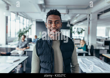 Portrait of happy young businessman standing in office with people sitting in background. African male designer standing in modern office. Stock Photo