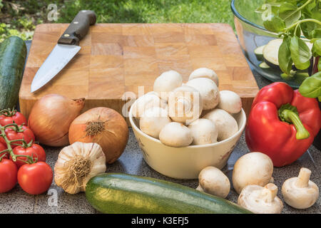 Wooden board, knife and various types of vegetables and mushrooms are lying on the table outdoors ready for food preparing. Stock Photo