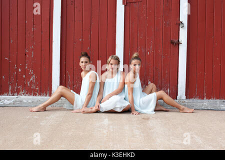 Three teenage sisters wearing matching white dresses sitting on the floor in front of a rustic red wall Stock Photo