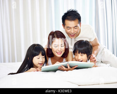 happy asian family with two children lying on front in bed reading a book together. Stock Photo