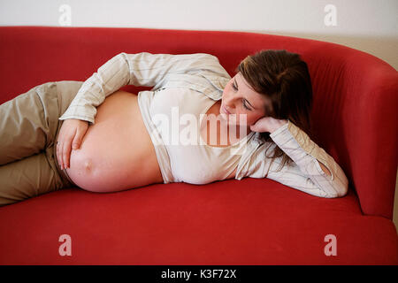 Pregnant young woman lies on red sofa Stock Photo
