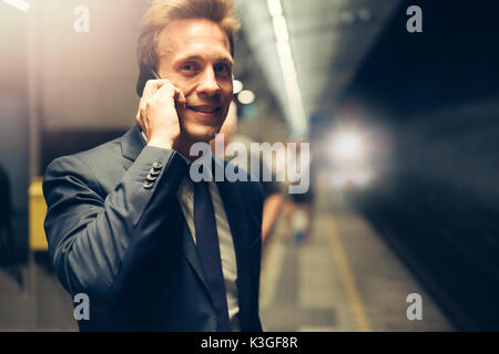 Smiling young businessman in a suit talking on his cellphone while standing on a subway platform during his morning commute
